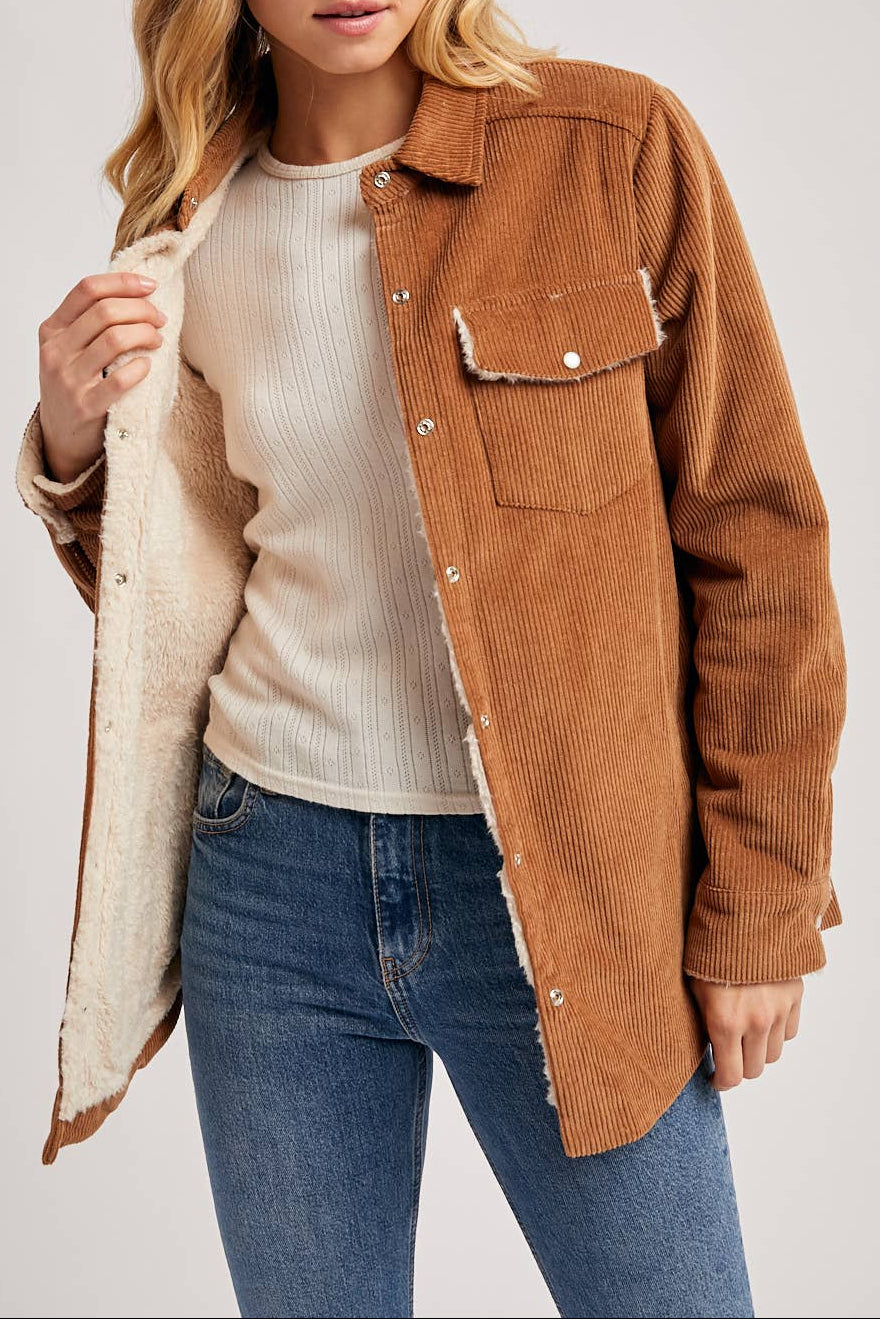 Woman wearing a brown corduroy jacket over a white shirt and blue jeans on a plain background