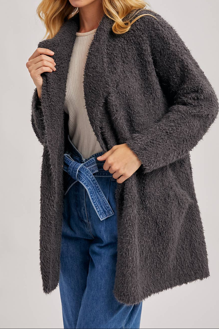 Woman wearing a dark gray fuzzy robe like cardigan over a light gray top and blue jeans on a plain background