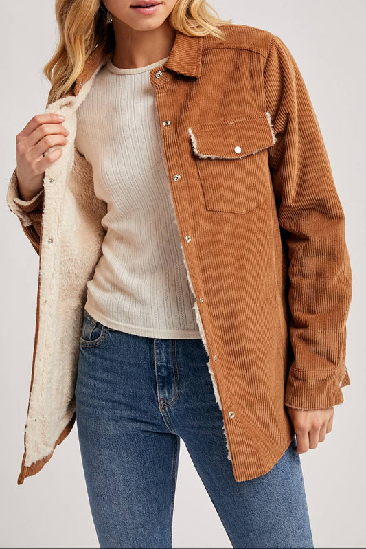 Woman wearing a brown corduroy jacket over a white shirt and blue jeans on a plain background